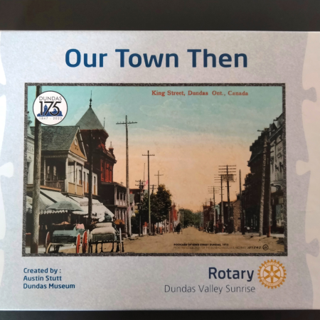 Image is of a cardboard box holding a puzzle. On the front it reads "our town then" and has the image of a lithograph postcard of King St., Dundas. It has the Dundas175 logo in the top left corner of the postcard.